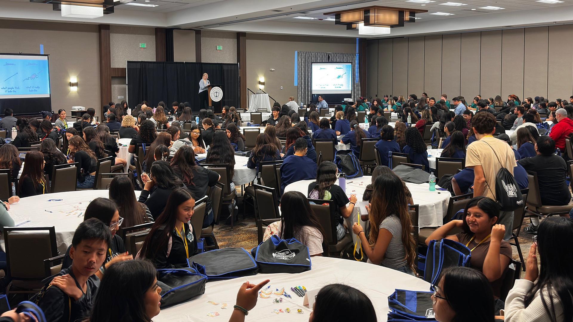 Students sit around many tables in a conference room, listening to a speaker.