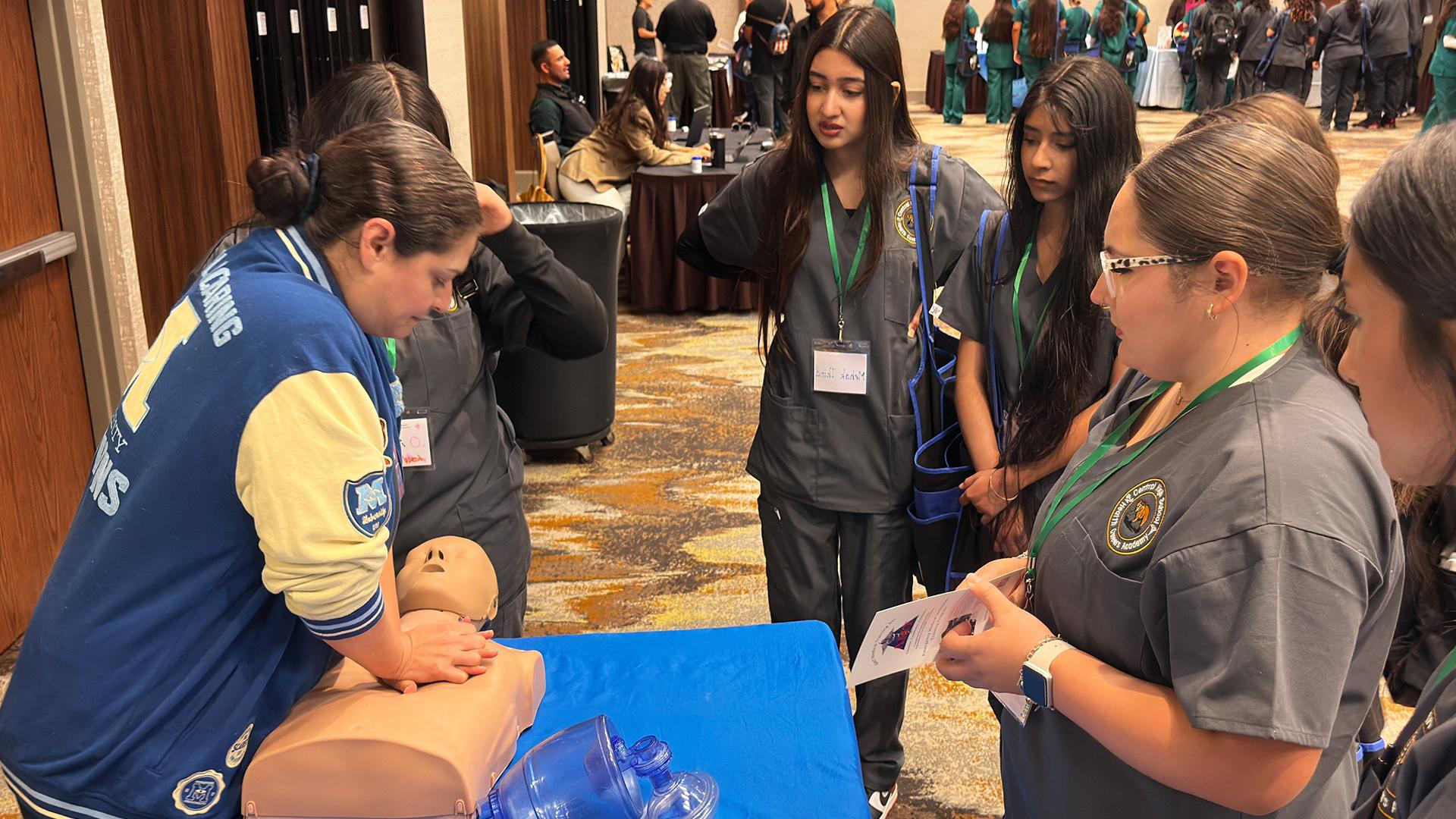 A group of students are gathered in front of a table watching a CPR demonstration.