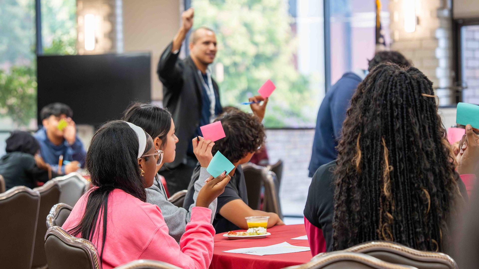 Students are sitting at tables, listening to a speaker while participating in a group exercise.