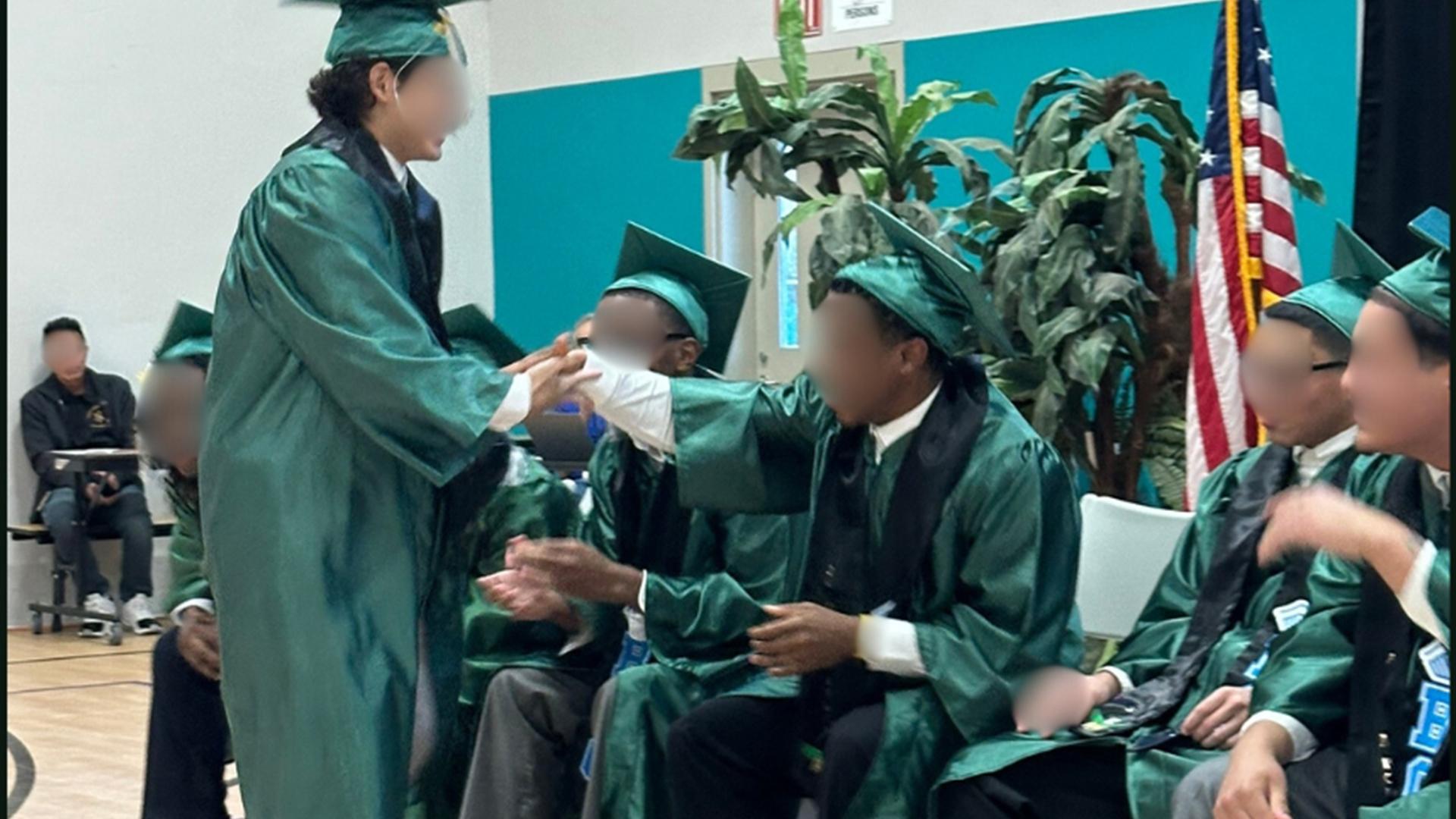 One graduate is standing, shaking hands with the other graduates who are sitting.