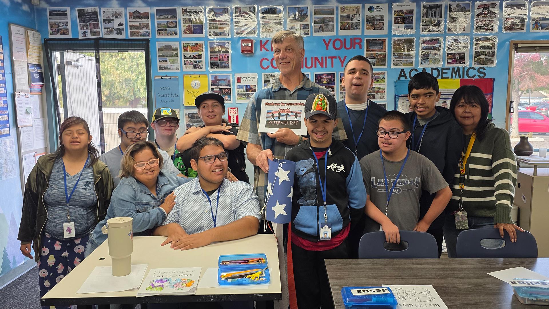 Smiling students are standing with their happy paraeducator with one student holding up the patriotic windsock they made for him.