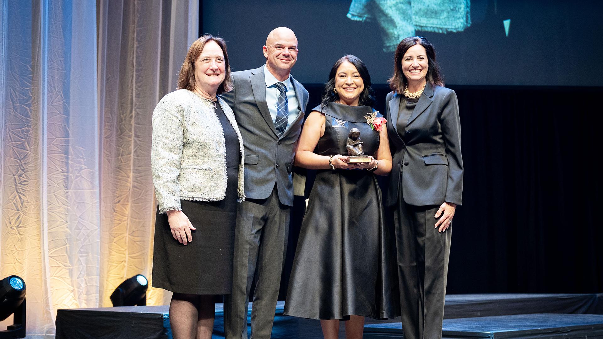 The Fresno County Teacher of the Year award winner is holding her award, smiling and flanked by the event sponsor and the Fresno County Superintendent of Schools.