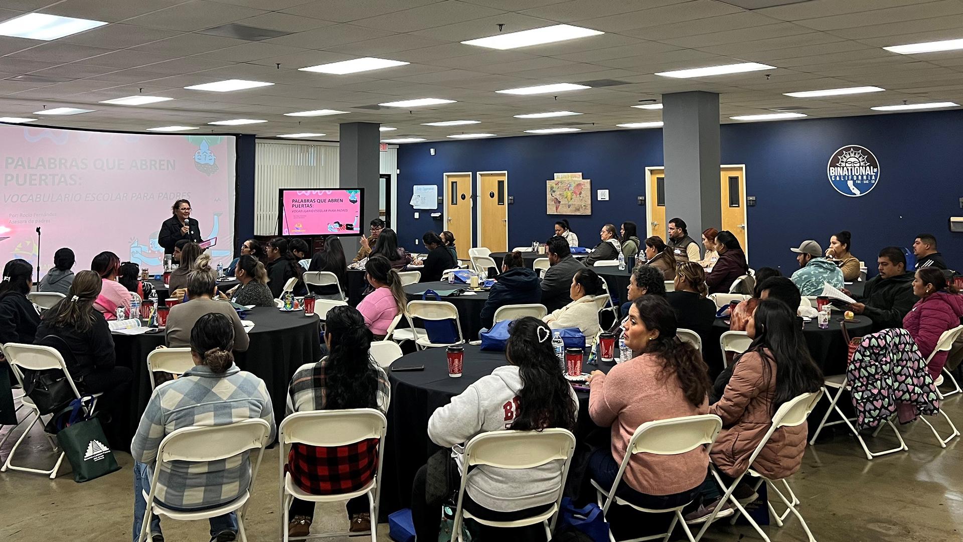 Parents and students are seated at tables, listening to a speaker.