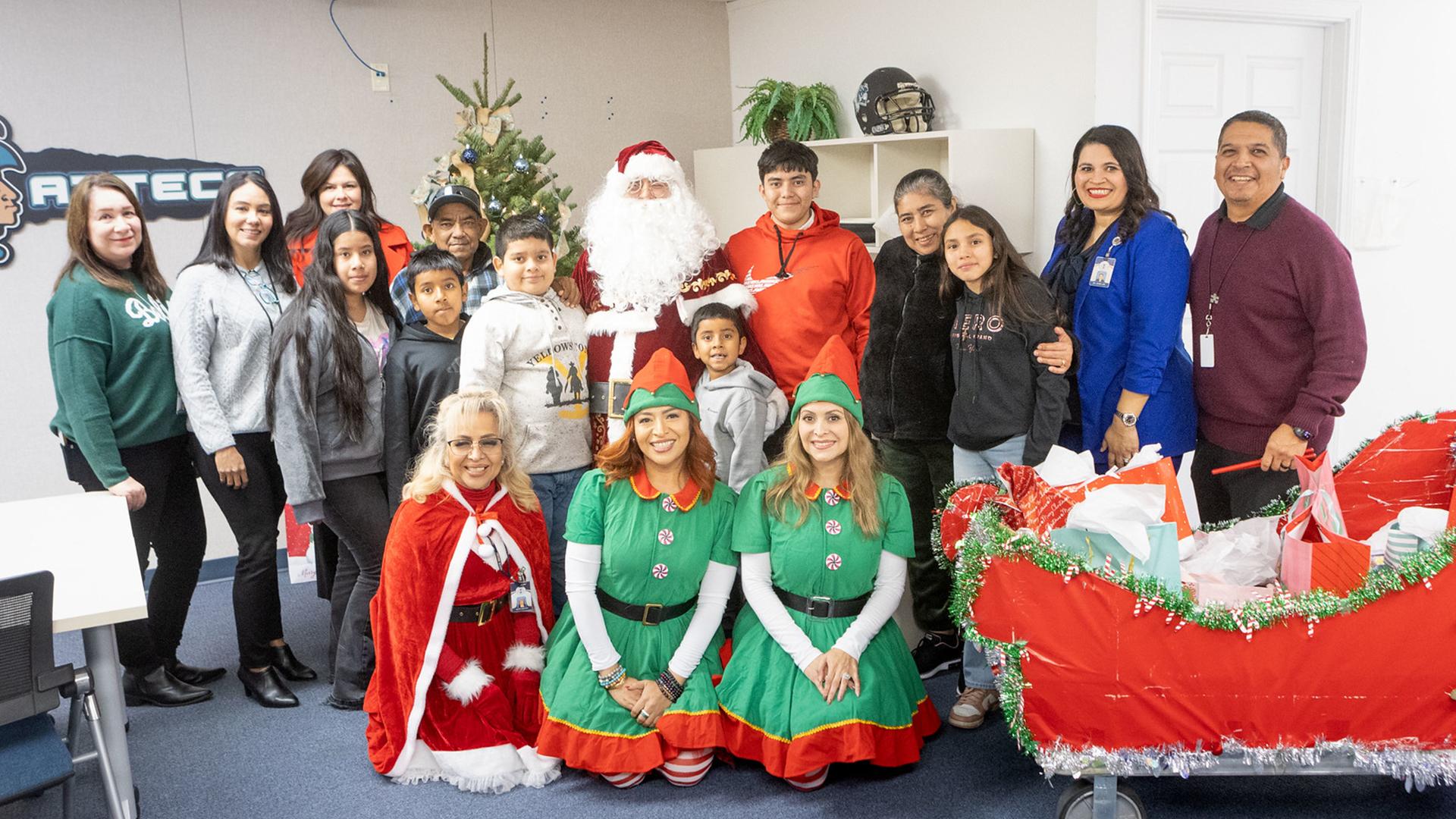 The family and staff are posed together, smiling, with Santa, elves and a sleigh full of gifts.
