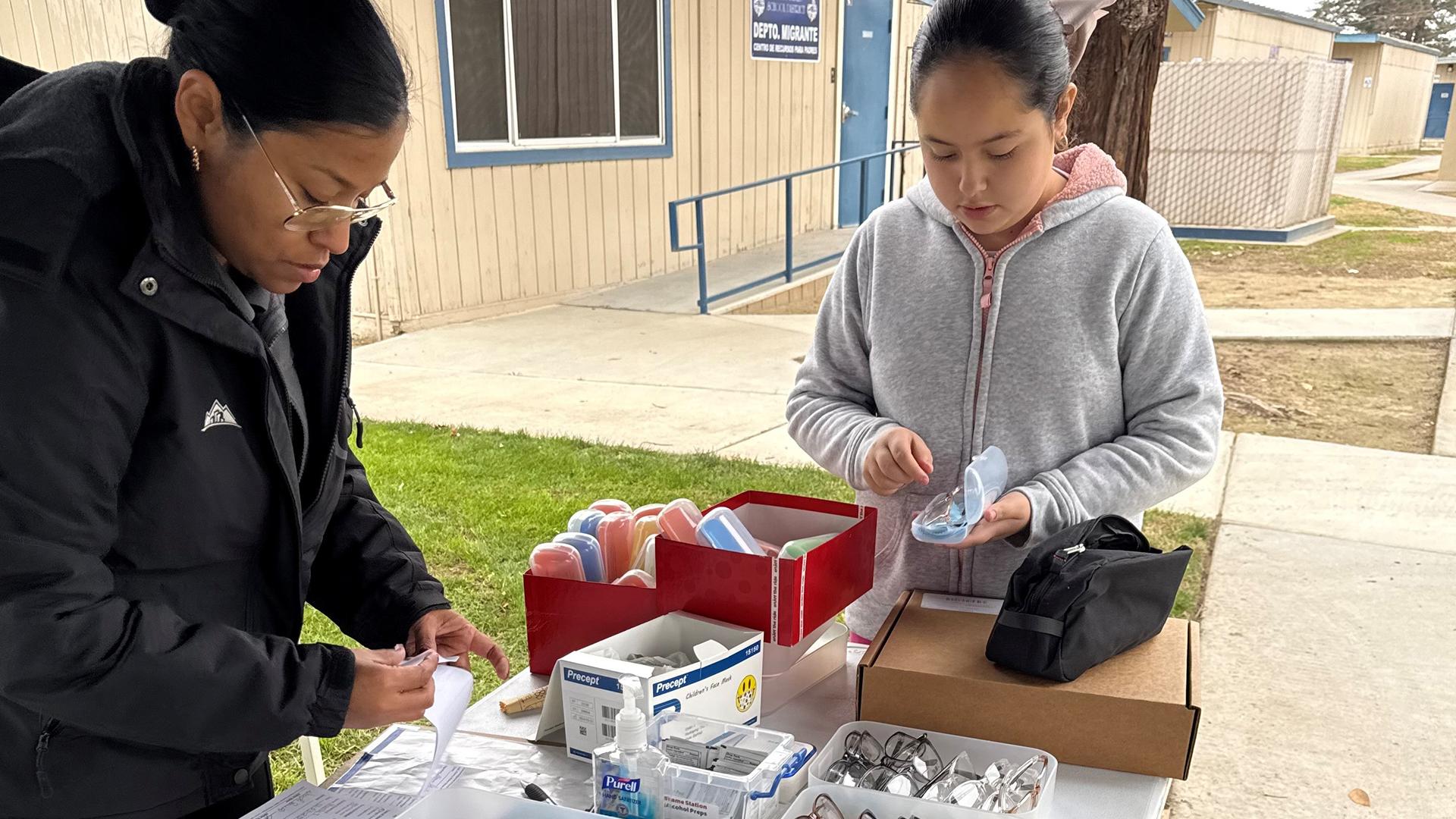 A student is standing with a staff member in front of a table full of glasses, looking at her own glasses in an open case.