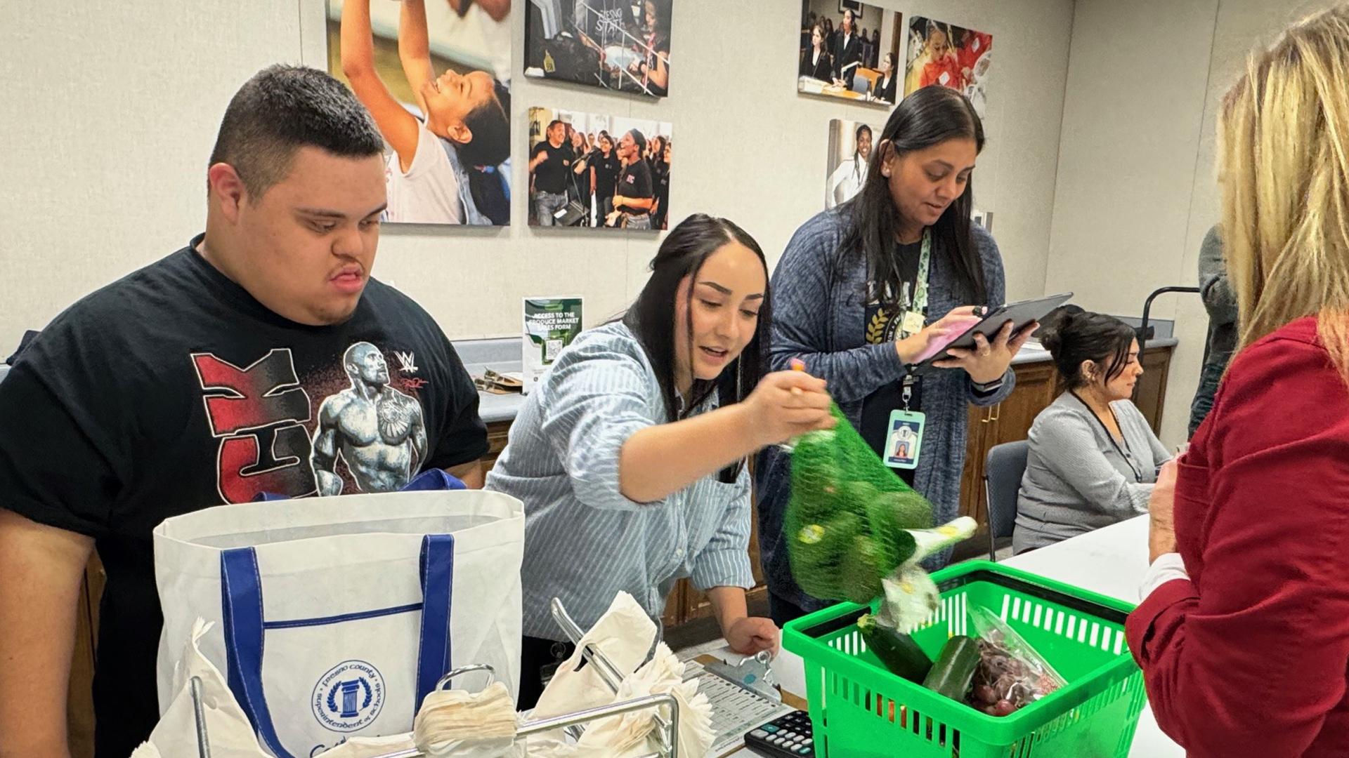 A staff member is purchasing produce at a table with a student and staff assisting her.