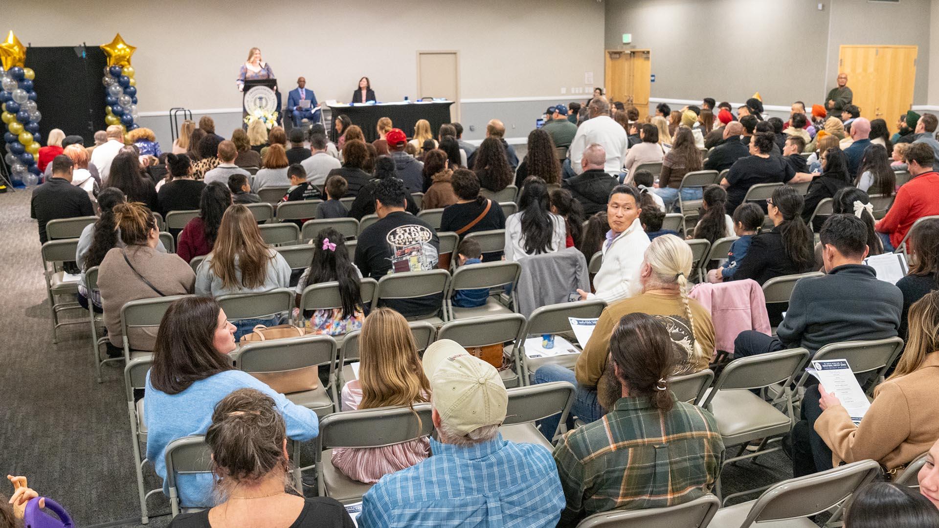 Families sitting in rows of chairs are listening to the awards ceremony presentation.