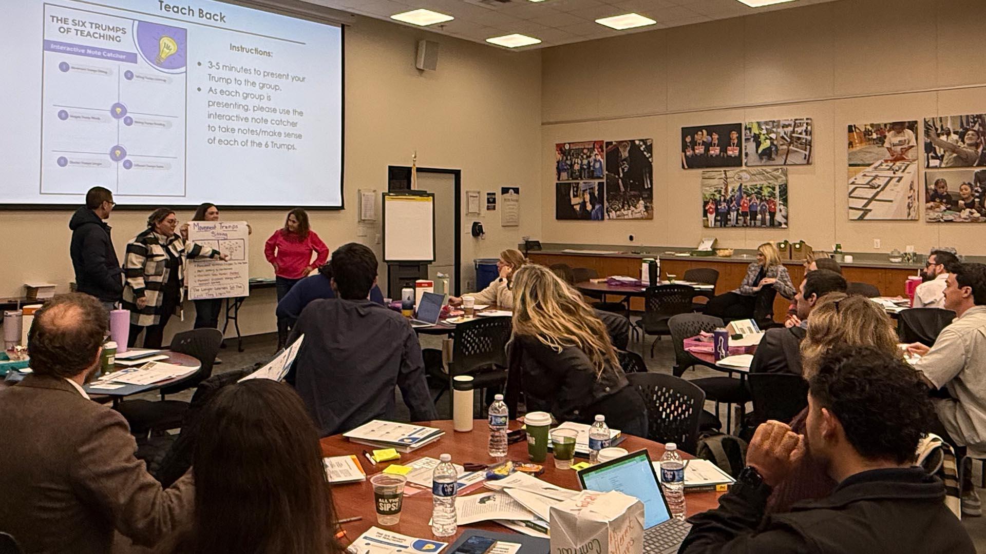 Workshop participants are seated at round tables, listening to the presenter.