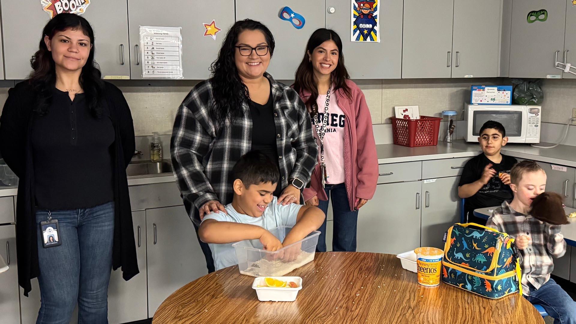 Students are sitting at tables, and staff are standing and smiling with them.