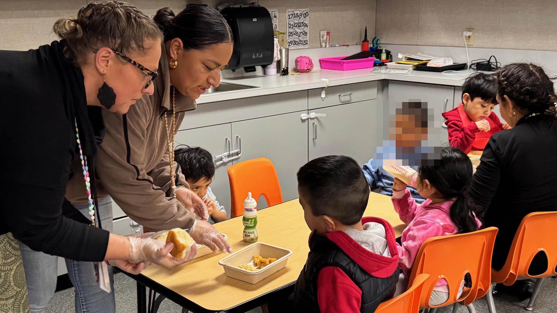 Teachers are helping their students eat breakfast at their classroom tables.