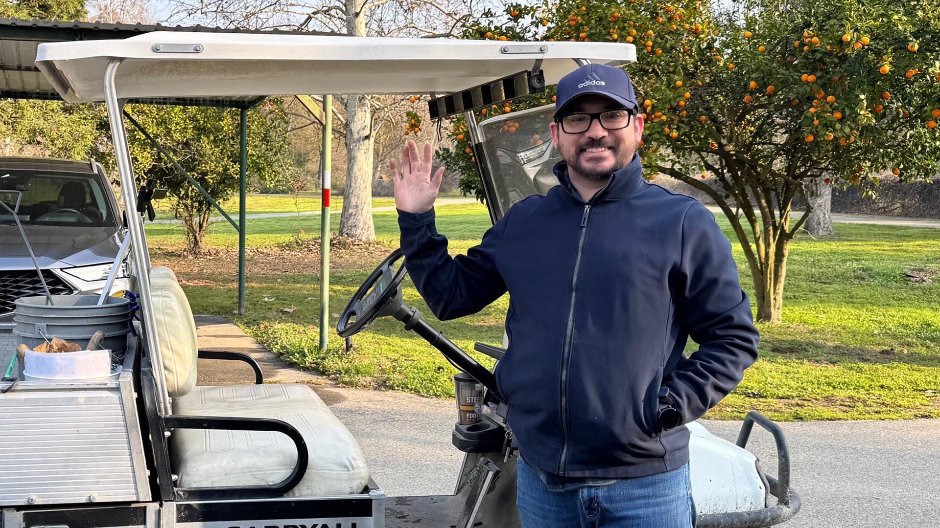 One person is standing, waving and smiling in front of a golf cart at Scout Island.