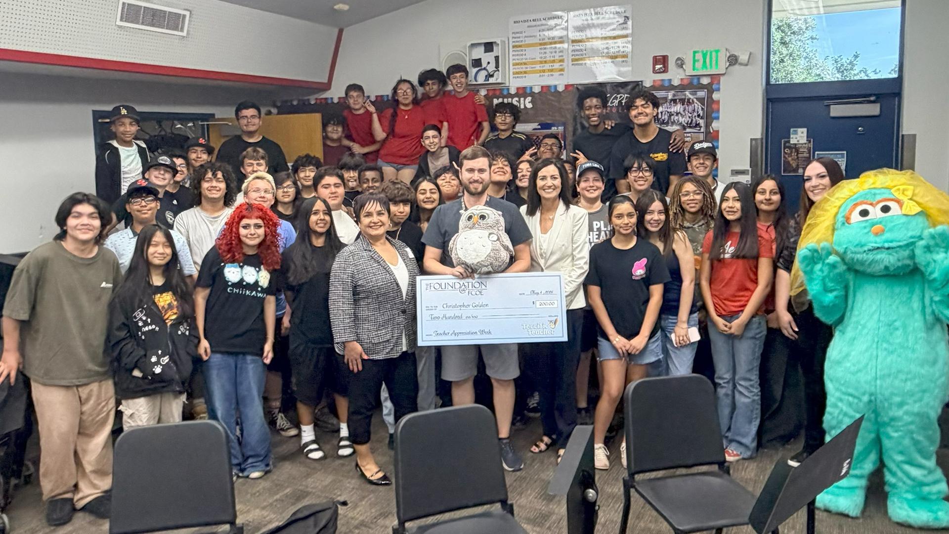 A classroom of students stand with a teacher, who receives an award. 