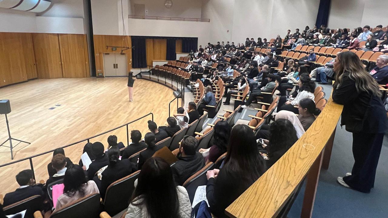 Students sit in an auditorium and listen to a speaker welcoming them to the event.